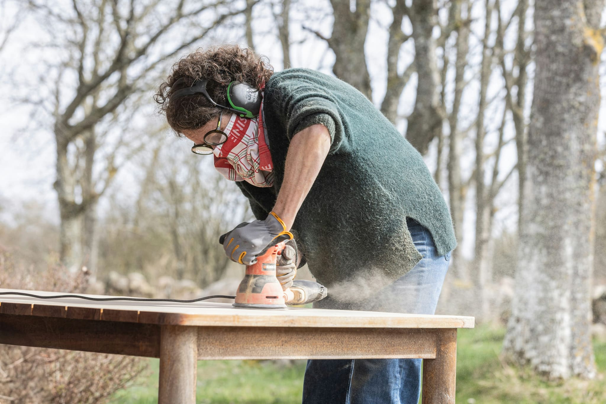 Symbolbild: Eine Frau mit Gehörschutz und Schutzbrille schleift im Freien einen Holztisch ab.